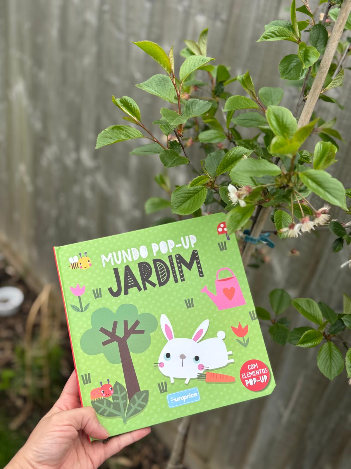 Children's pop-up book titled 'Mundo Pop-Up Jardim' held in front of a plant.