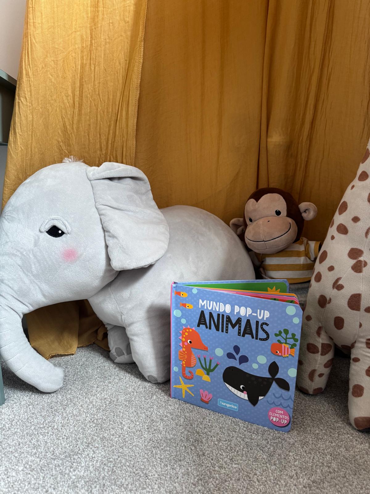 Children's book titled 'Mundo Pop-up Animais' surrounded by plush toys on a carpeted floor.