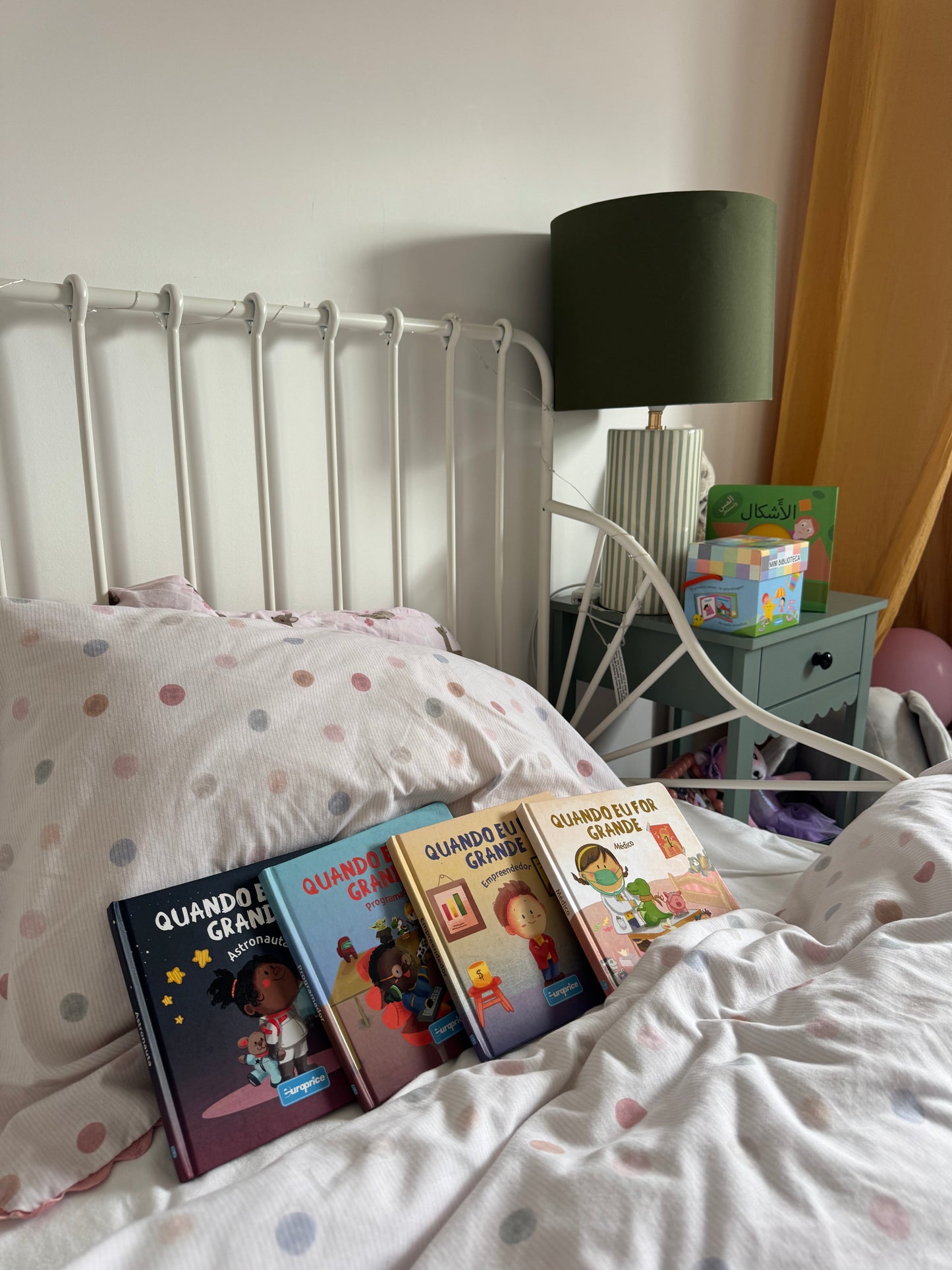Bed with polka dot bedding and children's books, nightstand with lamp and books in the background.